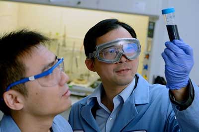 A man wears safety goggles and holds up a test tube with dark liquid and examines it while a second man observes