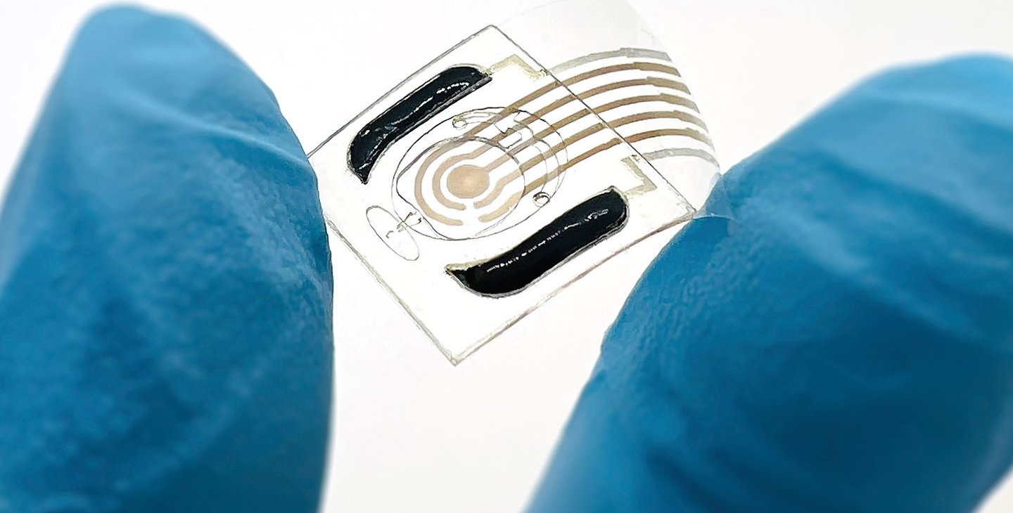 A hand wearing a blue rubber glove holds a semi-transparent plastic film containing a sensor between its index finger and thumb