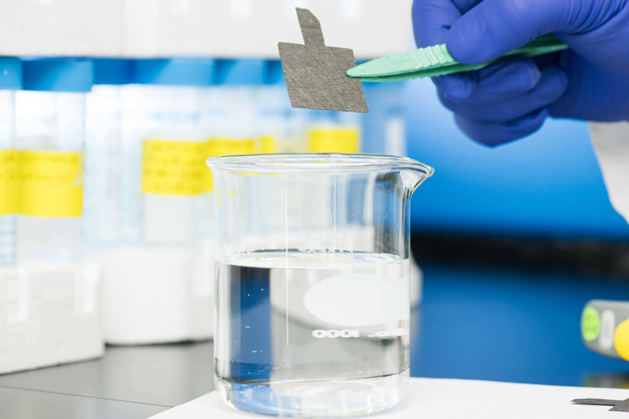 A gloved hands uses tweezers to hold carbon paper above a glass beaker filled with water to illustrate a new technique to remediate PFAS chemicals