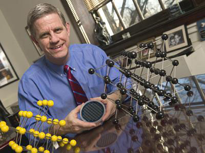Mechanical engineer Kevin Hemker, seated between models representing how atoms are packed within an individual grain in a material, holds a silicon wafer onto which nanocrystalline aluminum thin film specimens have been deposited.