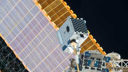 An image of NICER on the exterior of the space station with one of the station’s solar panels in the background